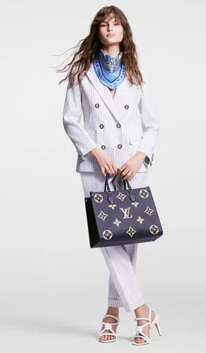 Model in pinstripe suit and blue scarf holding a dark monogram tote bag against a white background for Louis Vuitton Cruise.