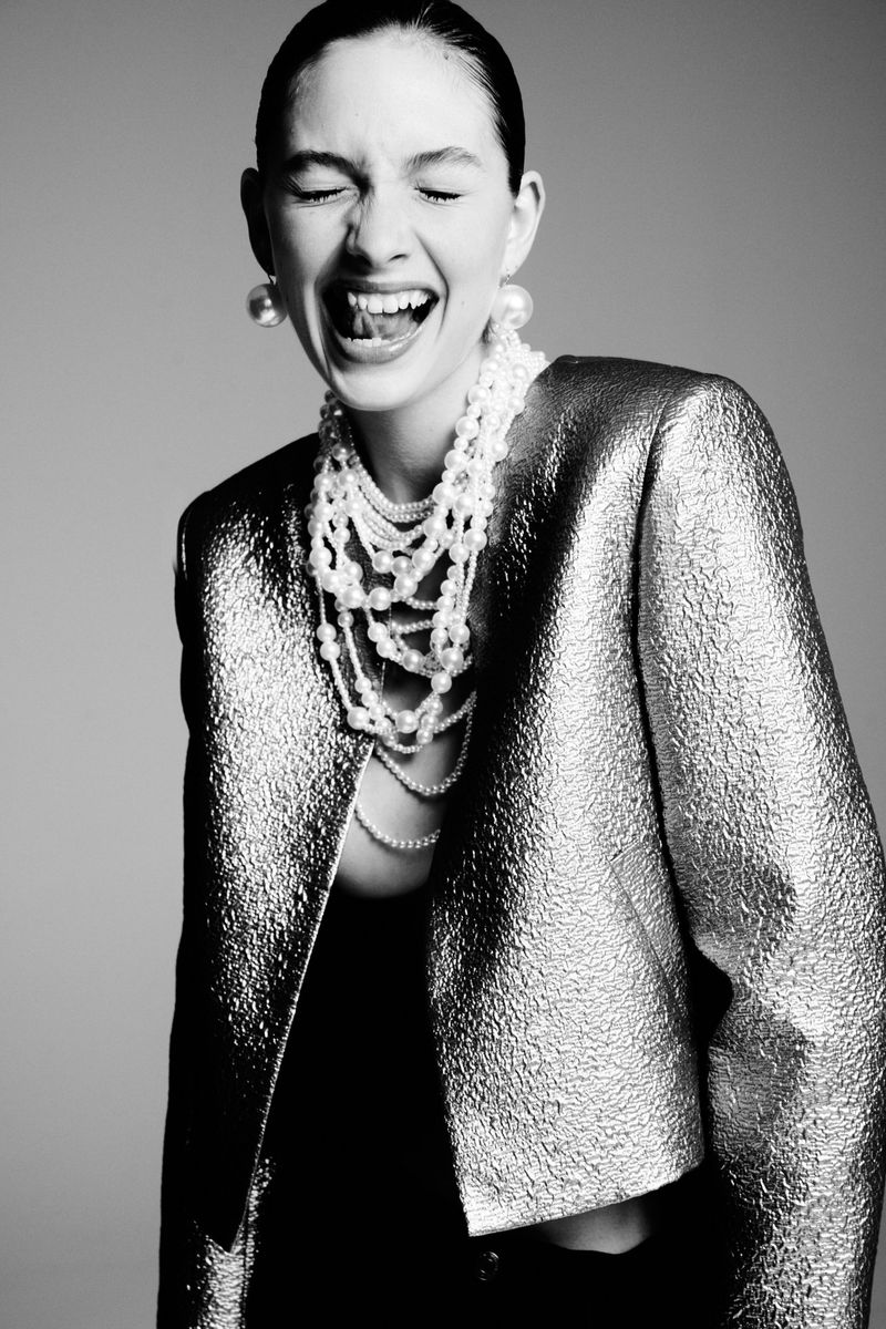 A black and white studio portrait captures a woman with an open mouth, wearing a metallic jacket and pearl jewelry. She is centered in the frame.