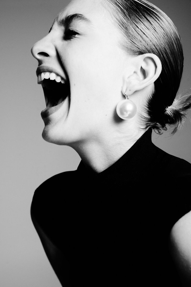 Black and white portrait of a woman with open mouth and pearl earring. Side-view of person wearing a dark top against a grey background.