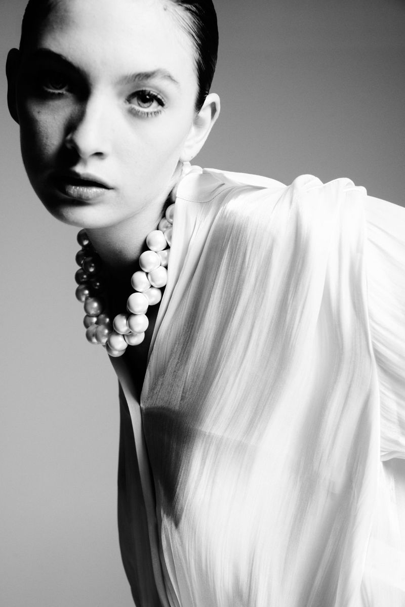 Monochrome portrait of a young woman with dark hair and a pearl necklace, wearing a draped white blouse against a gray backdrop.