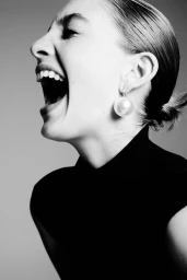 Profile portrait of a laughing woman wearing a large pearl earring and dark top against a grey seamless background