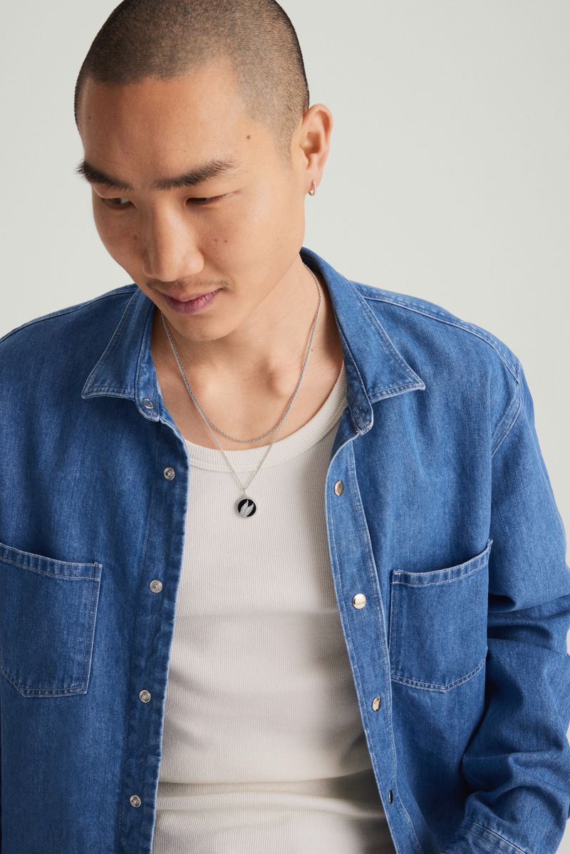 Close-up portrait of an Asian man wearing a blue denim shirt and silver necklace, looking down in a neutral studio setting.