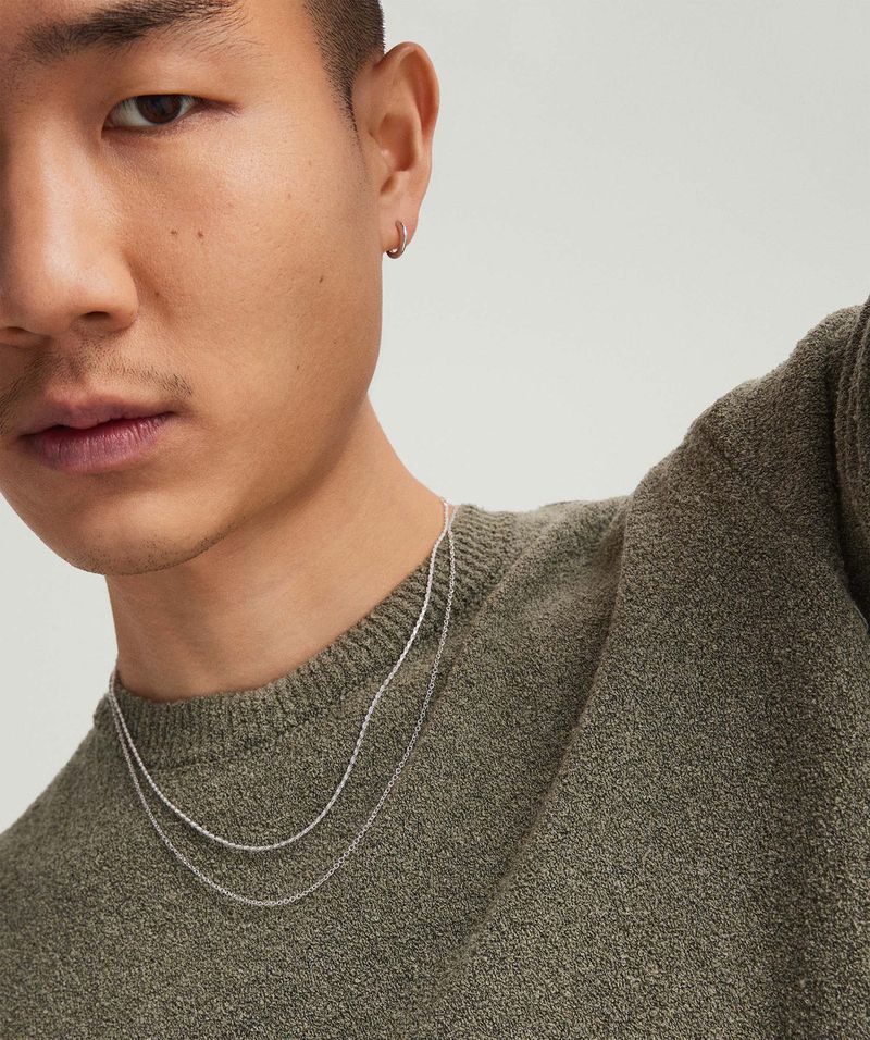 Close-up studio shot of an Asian man wearing a dark green sweater and layered silver chains. Fashion and beauty advertisement.