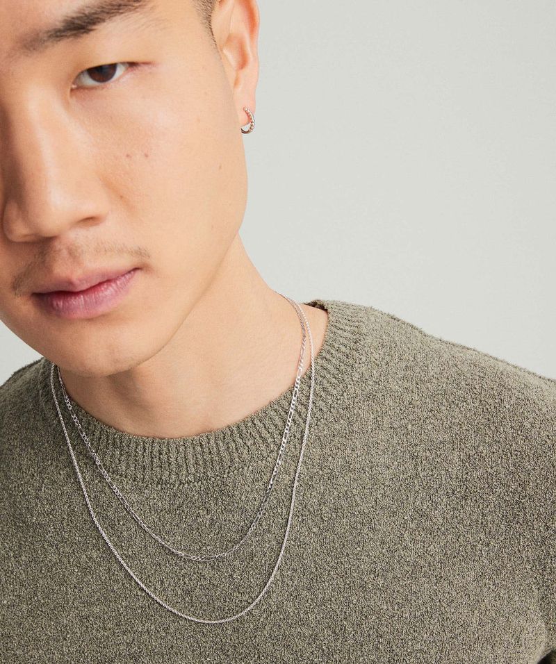 Close-up studio shot of a young adult Asian man wearing a knit sweater and layered silver chain necklaces; neutral background.