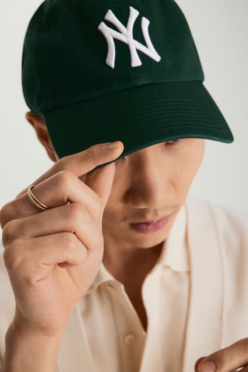 Close-up of an adult male wearing a dark green baseball cap with a white New York Yankees logo, gold ring, and a light tan button-down shirt.