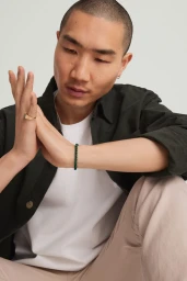 Close-up portrait of a man wearing a dark green shirt over white t-shirt, gold ring, and beaded bracelet against a pale background.