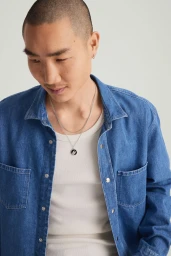 Close-up portrait of a man wearing layered silver necklaces, a white tank top, and a blue denim shirt against a pale background