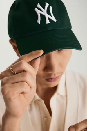 Close-up portrait of a man wearing a dark green baseball cap and gold rings on his fingers against a neutral light background.