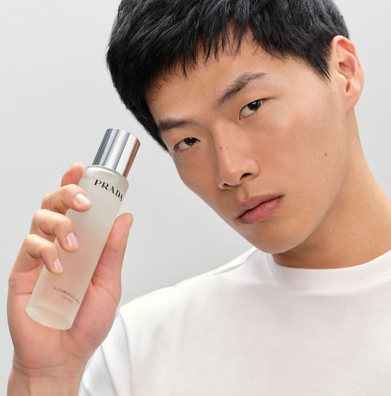 Close-up of an Asian male model holding Prada beauty serum, wearing a white T-shirt against a light background in a beauty product shot.