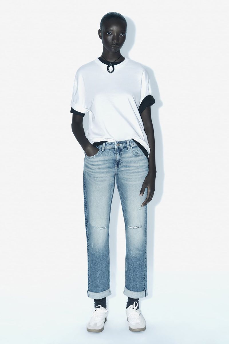 Full studio shot of an African-descent woman in a white t-shirt, distressed denim jeans, and tennis shoes against a white backdrop.