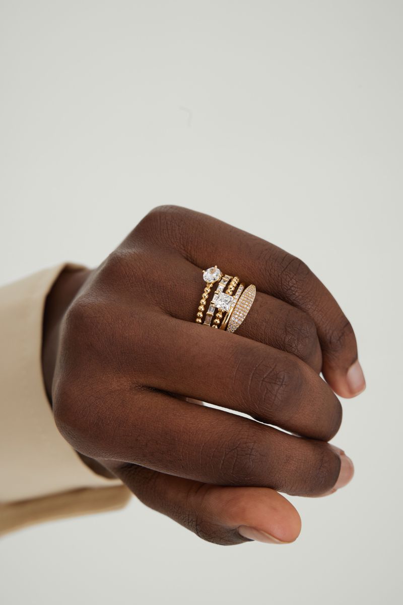 Close-up of a dark-skinned hand displaying an assortment of gold rings with clear gemstones against a neutral backdrop, studio lighting.