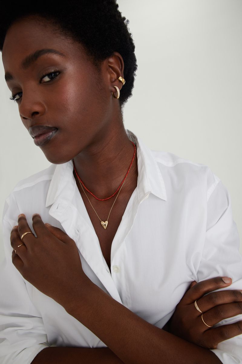 Close-up of a Black woman wearing a white button-down shirt, gold jewelry, and a red necklace against a neutral backdrop in a studio setting.