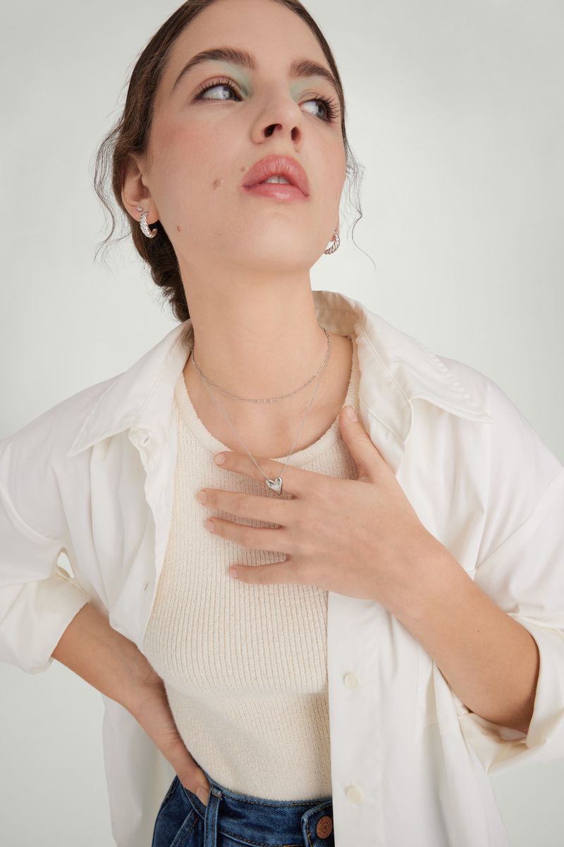 Portrait of a young adult woman with light skin wearing a casual white shirt, jeans, jewelry and light makeup, posing in a bright studio.
