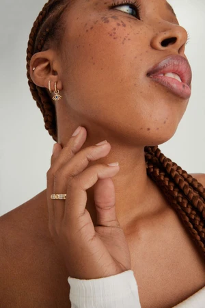 Close-up portrait of a woman wearing gold hoop earrings and a ring, with braided hair against a white background.