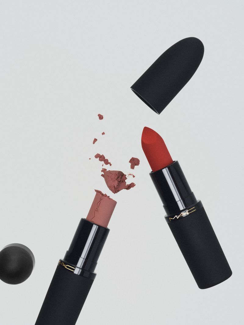 Two MAC lipsticks, one red and one neutral, with caps off and arranged diagonally on a light grey background in a beauty product shot.