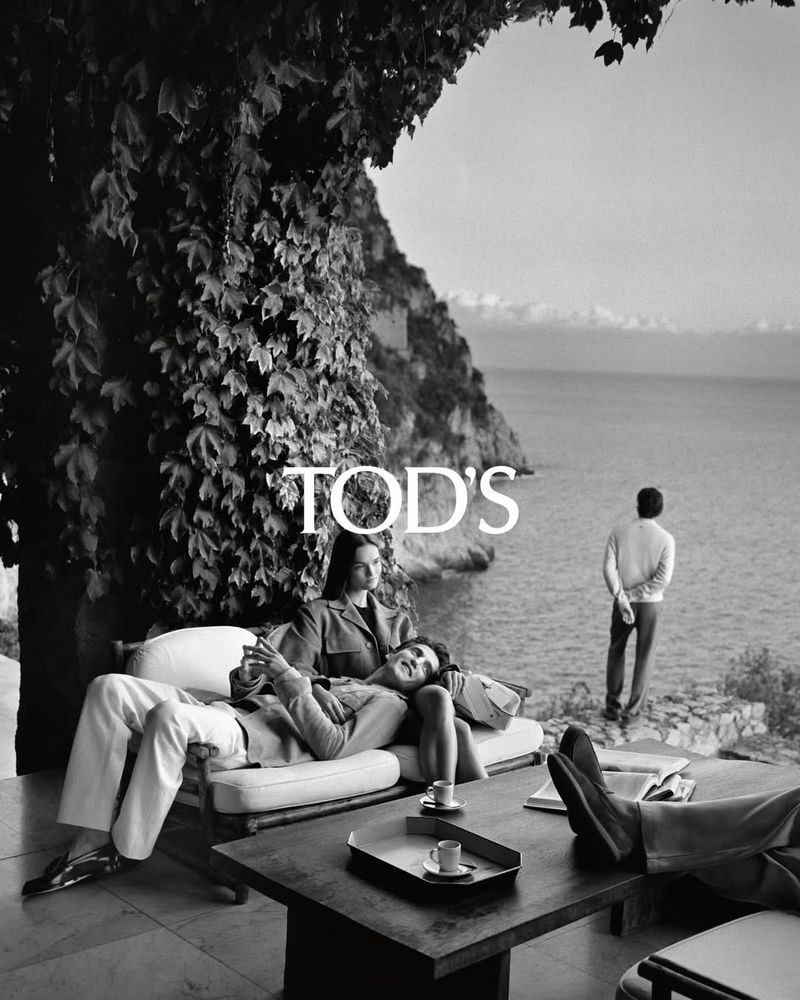 Black and white fashion shot featuring a couple lounging near the coast, a man standing nearby, and Tod's logo in the frame.