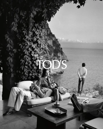 Black and white fashion shot featuring a couple lounging near the coast, a man standing nearby, and Tod's logo in the frame.