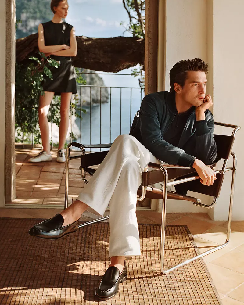 Man in white trousers and dark loafers sitting in a leather chair while a woman stands on a sunny balcony overlooking the sea.