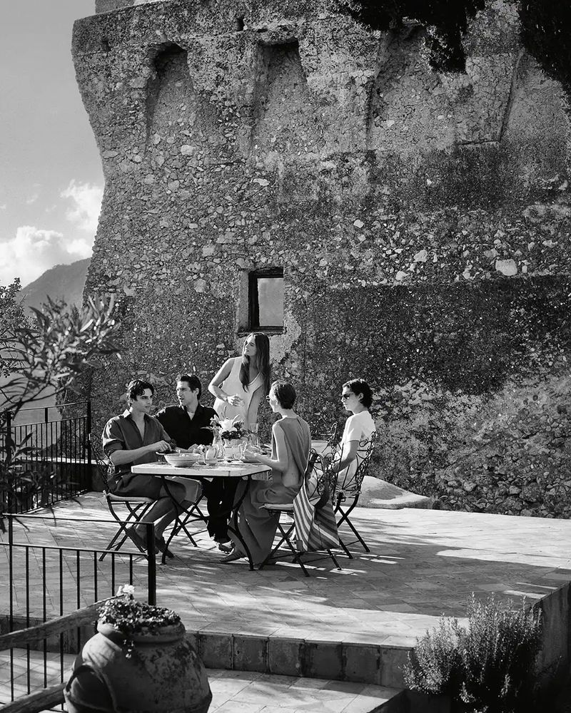Black and white image of five people at an outdoor table near a stone tower, enjoying a meal with drinks under natural light.