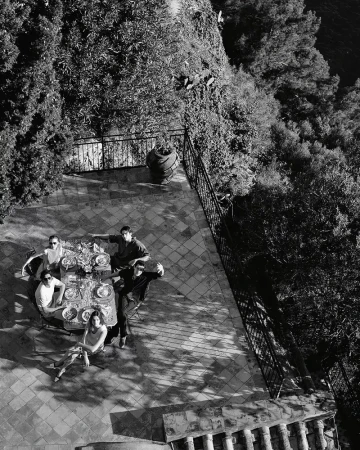 Aerial shot of six adults gathered around a table on a terrace lined with greenery and foliage on a sunny day, black and white.