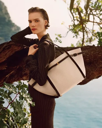 A light skinned woman poses outdoors with a large tote bag, wearing a dark, ribbed sweater dress, next to branches and a lake background.