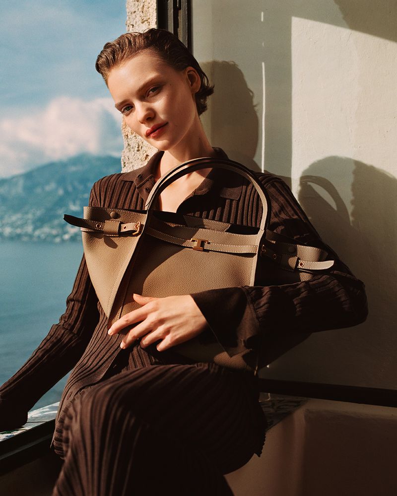 Attractive woman poses with a handbag in front of a scenic mountain lake, wearing a brown dress and natural makeup in soft lighting.
