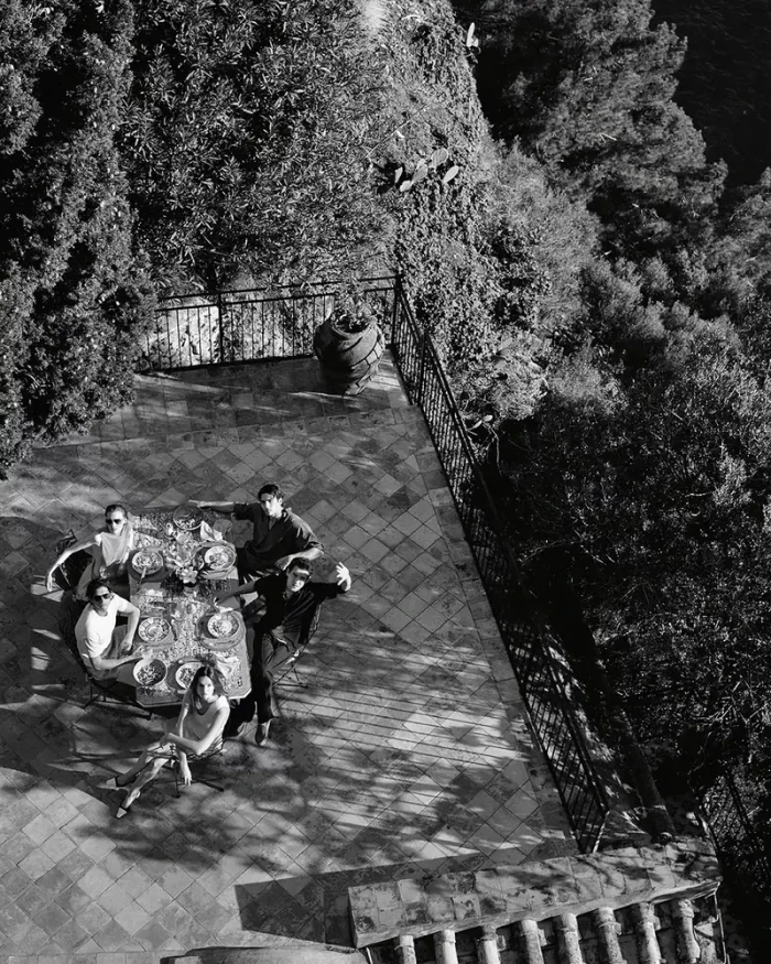 High-angle black and white shot of five people seated around an outdoor dining table on a tiled terrace bordered by lush trees.
