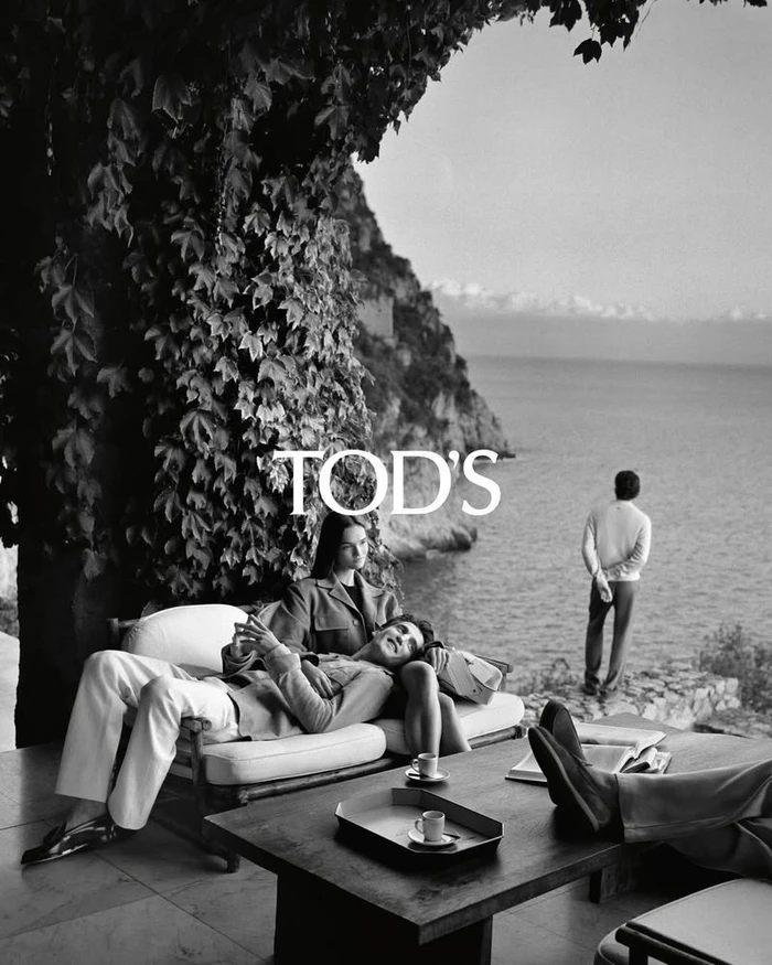 Black and white fashion campaign photograph featuring models lounging on a cliffside terrace overlooking the sea for Tod's.