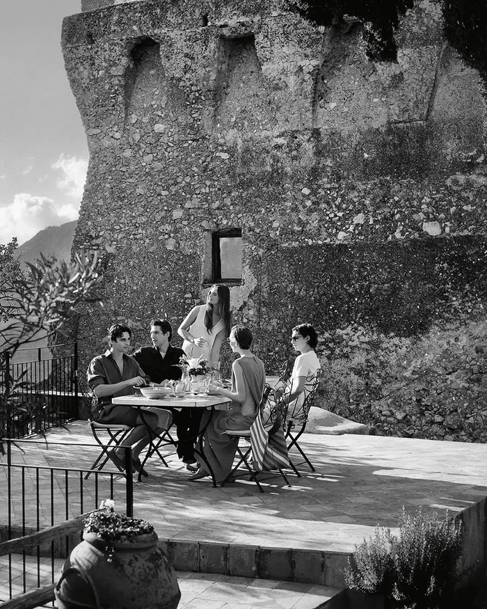 A group of five people seated and standing around an outdoor dining table beside a textured stone wall in black and white.