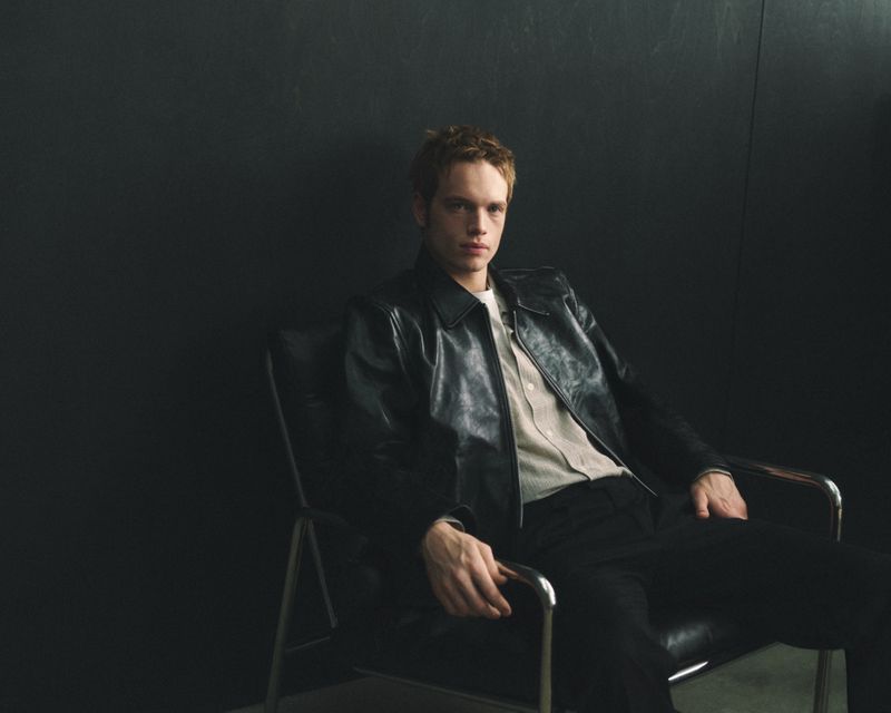 Man seated in a leather armchair wearing a black leather jacket against a dark wall for Mr Porter Essentials Campaign