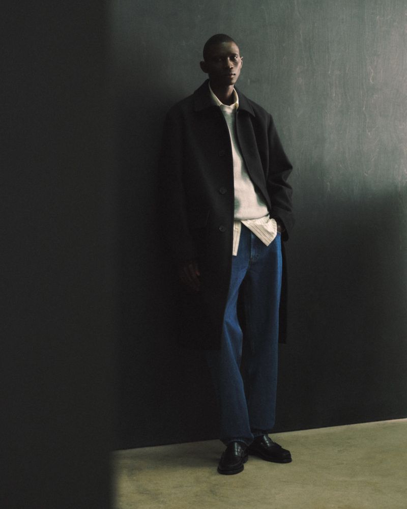 Dark-skinned male model in jeans and a coat stands against a dark wall, exuding a modern, urban fashion aesthetic in studio lighting.