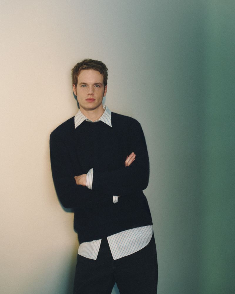 Man in a black crewneck sweater over a striped button-down shirt leaning against a gradient wall with arms crossed.