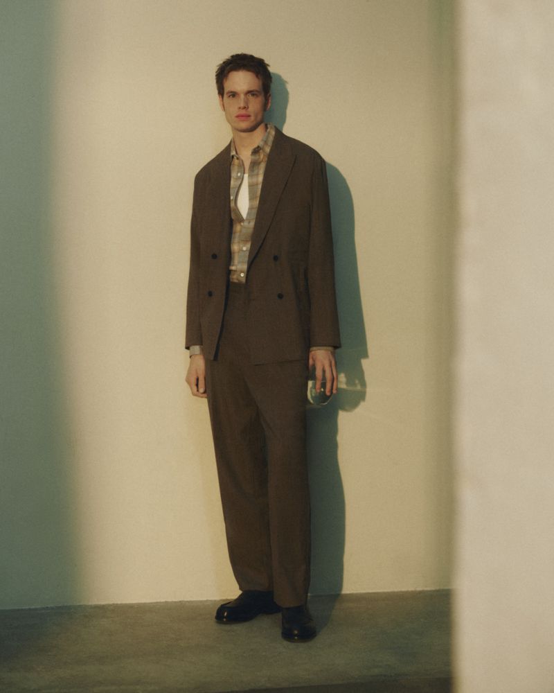 A full-length studio shot of a young adult male wearing a brown suit and patterned shirt, holding a glass, against a neutral wall.