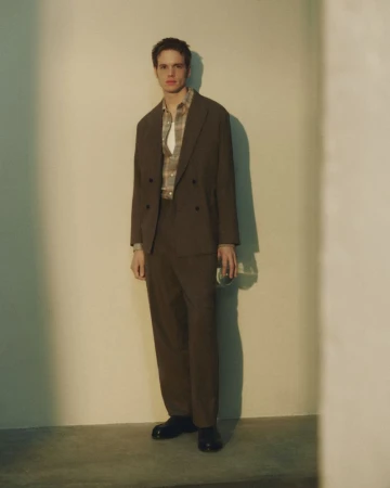 A full-length studio shot of a young adult male wearing a brown suit and patterned shirt, holding a glass, against a neutral wall.