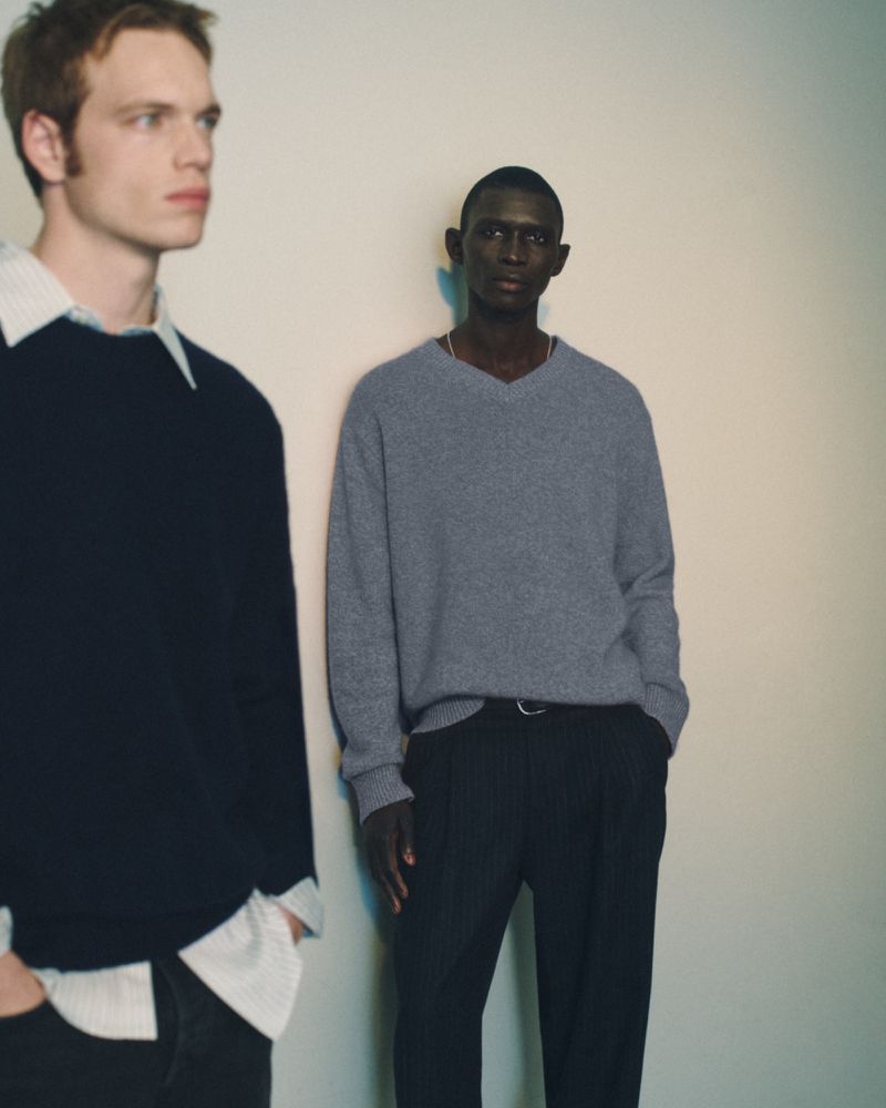 Two men in sweaters pose indoors against a neutral wall. One is fair-skinned with an open collar, while the other has darker skin.