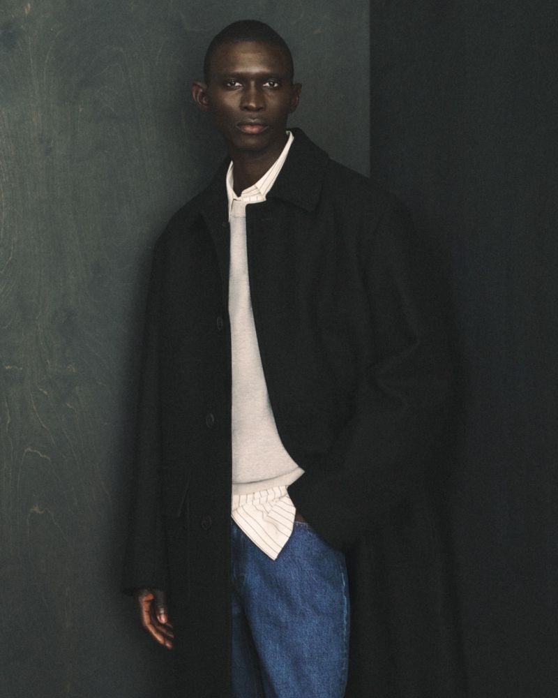 An adult Black male in a coat and jeans poses against a dark gray corner in a fashion portrait with eye-level angle and soft lighting.