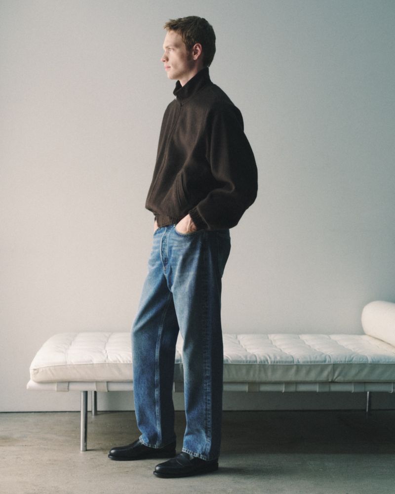 An adult male stands indoors with hands in his denim jeans pockets, wearing a dark brown sweater and black shoes next to a daybed.