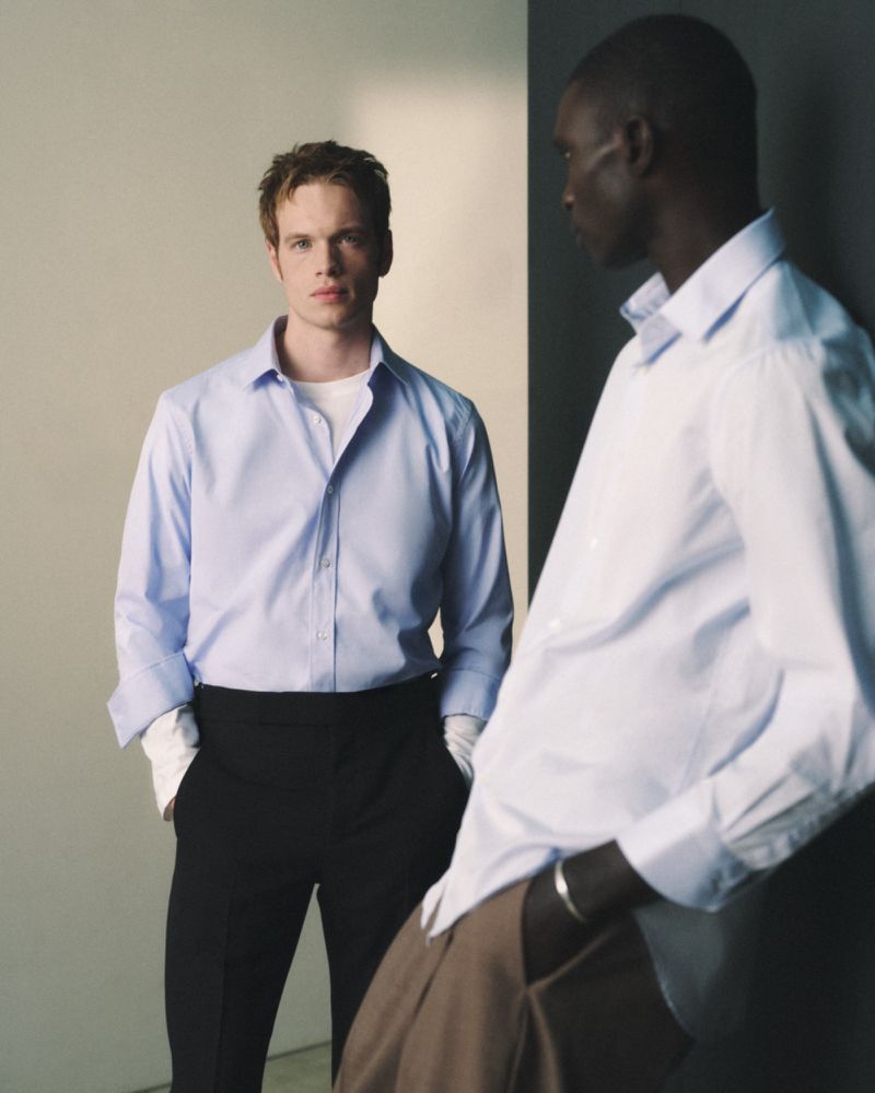 Two men pose in a minimalist studio wearing collared shirts and dark pants against a neutral background with a split-tone color scheme.
