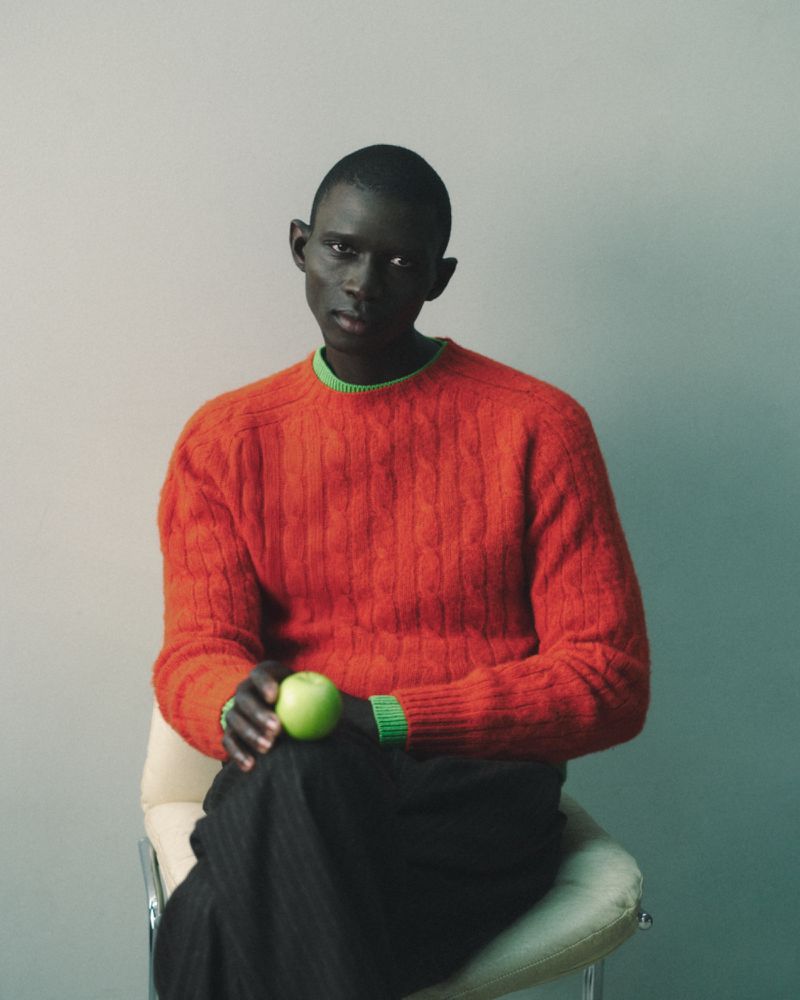 Portrait of an adult male of African descent sitting on a chair holding a green apple, wearing an orange sweater and black pants in studio.