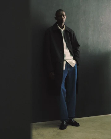 Model Fernando Cabral posing against a dark wall in a black overcoat, cream knit sweater, blue denim jeans, and black loafers.