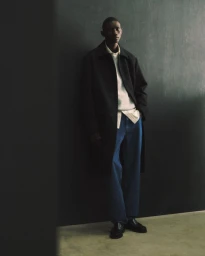 Model Fernando Cabral posing against a dark wall in a black overcoat, cream knit sweater, blue denim jeans, and black loafers.
