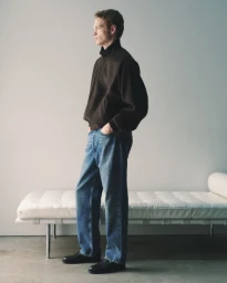 Model standing in a dark zip jacket, relaxed blue jeans, and black loafers beside a white daybed in a studio.