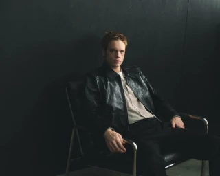 Man seated in a leather armchair wearing a black leather jacket against a dark wall for Mr Porter Essentials Campaign