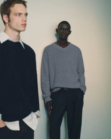 Fernando Cabral wearing a grey V-neck sweater and black trousers alongside another model in a dark sweater against a pale studio wall.