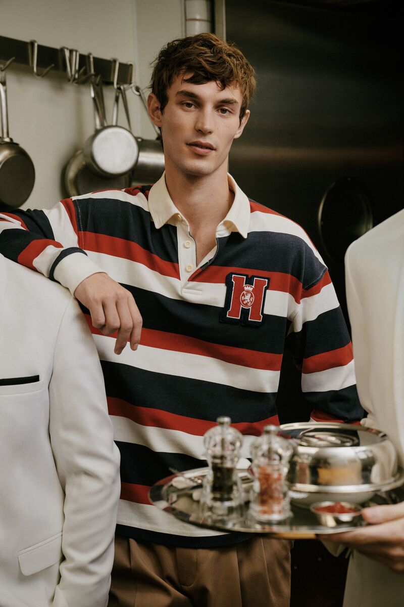 Man in a striped rugby shirt and tan trousers stands next to a waiter holding a silver tray in an indoor setting.