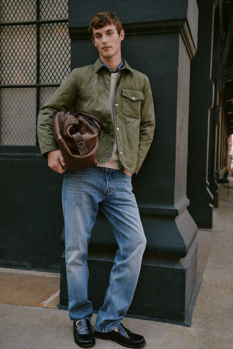 A stylish adult male model in a quilted jacket, jeans, and loafers poses with a brown leather duffel bag near a building.