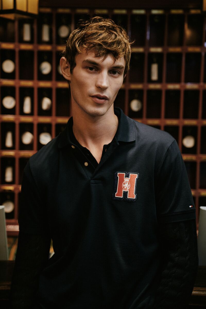 Headshot of fair-skinned male model in black polo shirt with 'H' logo against a blurred background of wooden compartments.