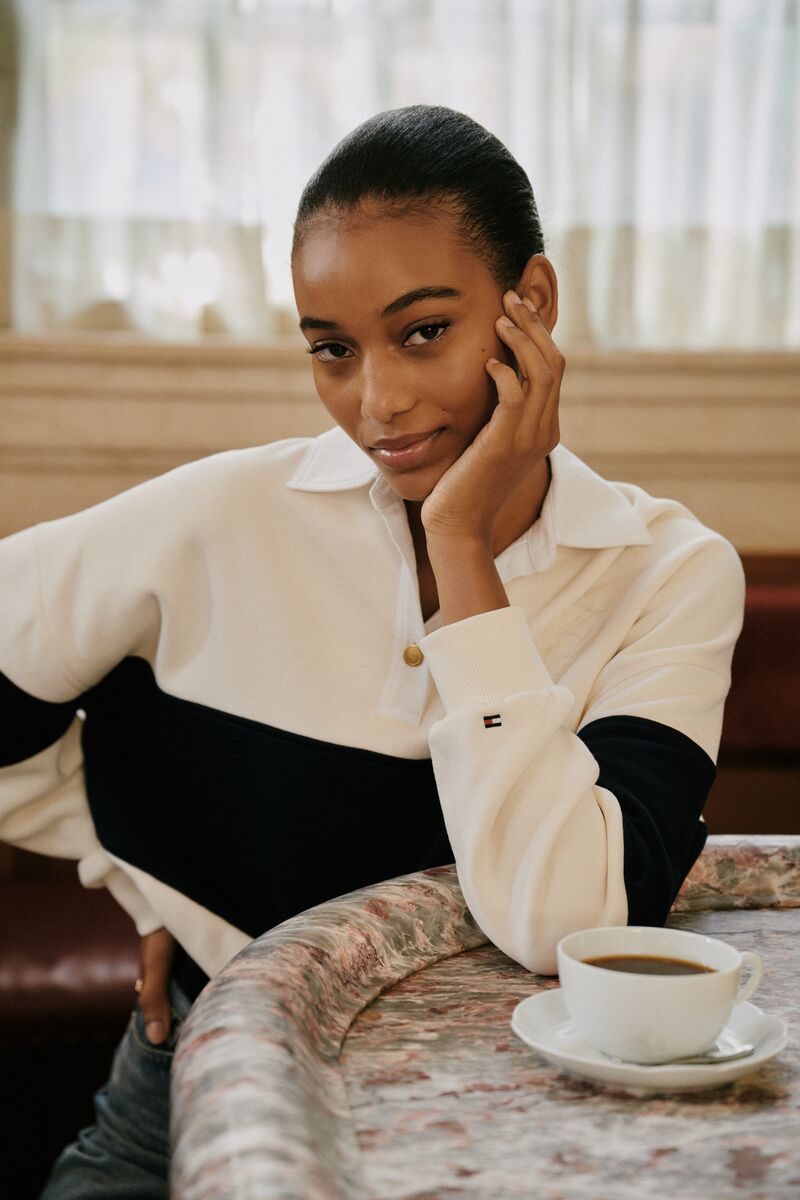 Portrait of a young Black woman with short hair wearing a cream and navy top, resting her head on her hand at a cafe table.
