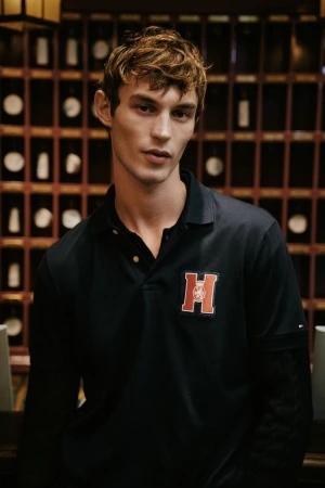 Close-up portrait of model Kit Butler wearing a navy polo shirt with an embroidered logo against a dark wood wall.