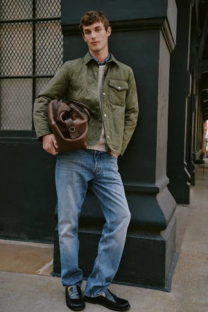 Model wearing an olive quilted jacket and blue jeans holding a brown leather duffel bag while leaning against a dark column.